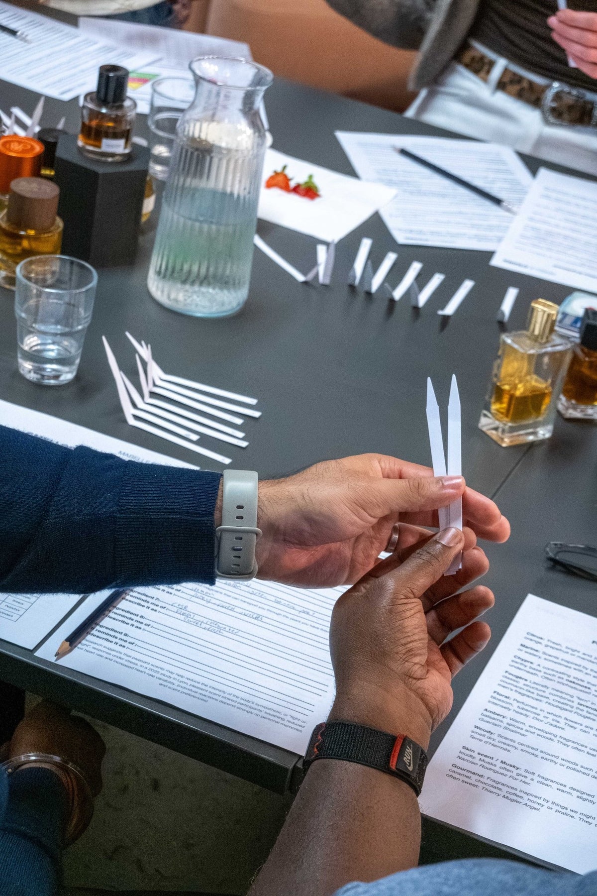Hands holding two scent strips over a table of perfumes and notes